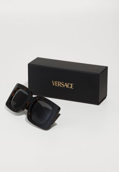 Melnas, pārāk lielas Versace saulesbrilles ar tumšiem objektīviem un bruņurupuča rakstu, novietotas blakus melnai kārbiņai ar zelta "VERSACE" tekstu.