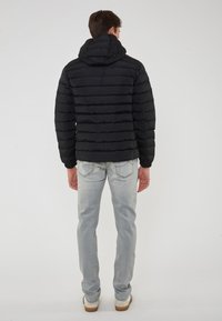 Giacca a vento nera con design a cuciture orizzontali, cappuccio attaccato e polsini elastici, abbinata a jeans grigi chiari e scarpe casual.