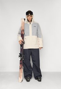 Strój snowboardowy składa się z beżowej i szarej kurtki z dużymi kieszeniami, w połączeniu z luźnymi ciemnymi spodniami, czarnymi butami i sprzętem narciarskim.