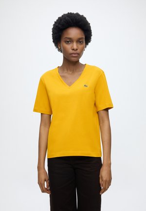 Femme aux cheveux courts et bouclés porte un T-shirt jaune à col en V et un pantalon noir, debout devant un fond clair uni.