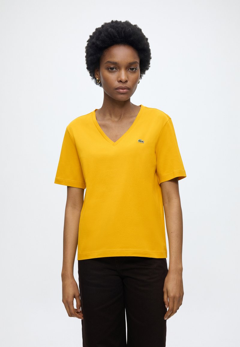 Femme aux cheveux courts et bouclés porte un T-shirt jaune à col en V et un pantalon noir, debout devant un fond clair uni.