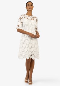 Weißes Spitzenkleid mit floralem Muster, kurzen Ärmeln und einem gewellten Saum. Kombiniert mit leichten Sandalen mit Riemchen.