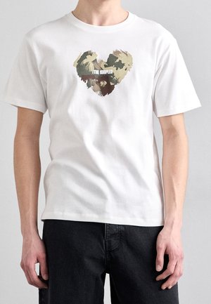 Homme portant un t-shirt blanc avec un cœur camouflage abstrait et le texte "THE KOOPLES", associé à un pantalon noir, sur un fond uni.