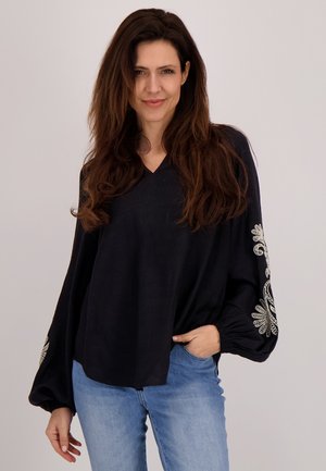 Zwarte blouse met lange mouwen en een V-hals, versierd met fraaie witte borduursels op de mouwen. Gepaard met lichtblauwe jeans in een relaxte pasvorm.