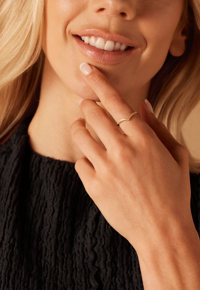 Gouden ring met een golvend ontwerp, gedragen aan een hand die op de kin steunt. De achtergrond heeft een textuur van zwart stof en een neutrale huidtint.