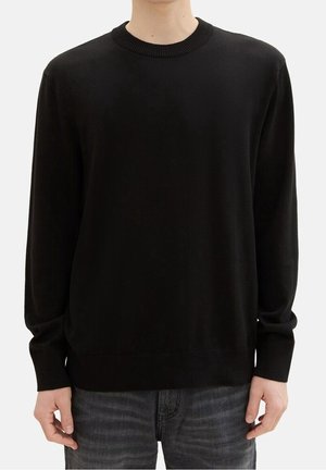 Personne portant un pull noir uni à manches longues et col rond, ainsi qu'un jean gris foncé, debout devant un fond blanc.