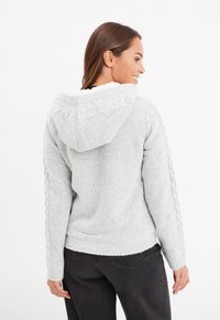 Felpa grigia con cappuccio in tessuto testurizzato, dettagli a treccia sulle maniche e cappuccio foderato in pile. Presenta una vestibilità casual e polsini a coste.