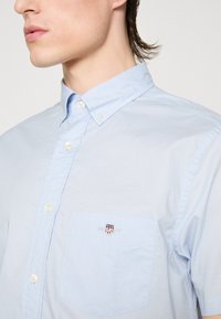 Homme portant une chemise bleu clair boutonnée avec un petit emblème en forme d'écu rouge, blanc et bleu sur la poche poitrine.
