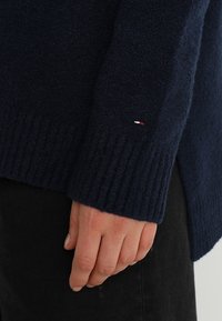 Mörkblå stickad tröja med ribbad ärmslut; har en liten detalj med röd, vit och blå logotyp nära ärmen. Mjuk textur.