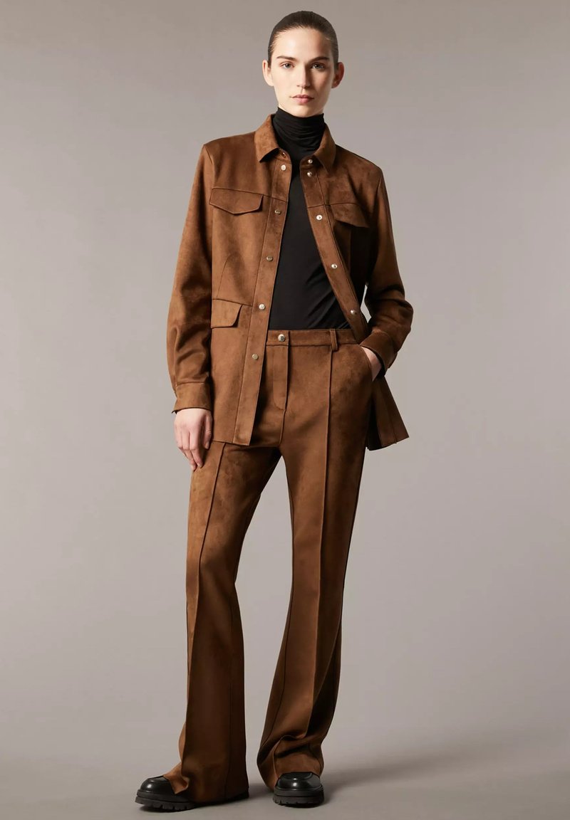 Giubbotto in suede marrone con tasche frontali, abbinato a pantaloni a vita alta coordinati. Presenta una finitura testurizzata e una maglia a collo alto nera sotto.