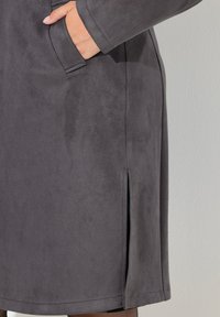 Abrigo de ante gris con un diseño recto, bolsillos laterales y discretas aberturas laterales en el dobladillo para facilitar el movimiento. Textura suave y líneas limpias.