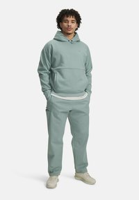 Svetlozelená mikina s kapucňou a klokanie vrecko, spárovaná s voľnými nohavicami v rovnakom odtieni a béžovými teniskami. Hladká látka, minimálne brandovanie.
