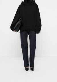 Sudadera negra oversize con capucha y logo, combinada con jeans azul oscuro de corte recto y tacones negros de punta afilada. Bolso de mano negro.