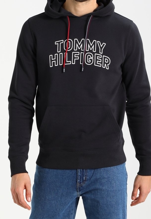 Sweat à capuche noir avec poche avant, logo "TOMMY HILFIGER" en blanc, cordons de serrage rouges et bleu marine, porté avec un jean bleu.