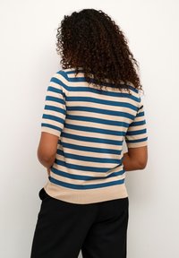 Kobieta z kręconymi włosami ubrana w niebiesko-beżowy sweter w paski z krótkim rękawem oraz czarne spodnie, stojąca plecami do białej, gładkiej ściany.