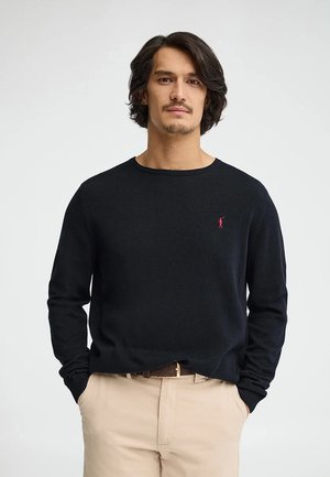 Uomo con capelli castani di media lunghezza che indossa un maglione nero con logo rosso e pantaloni beige, in piedi con le mani nelle tasche.