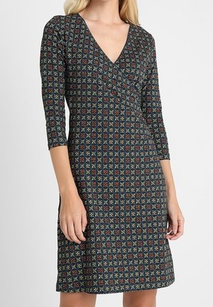 Vrouw draagt een knielange jurk met driekwart mouwen en een geometrisch patroon in donkere kleuren met rode accenten.