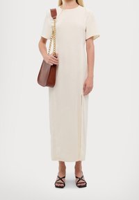 Robe midi couleur crème avec manches courtes, présentant une fente latérale. Le modèle tient un sac en cuir marron avec une bandoulière chaîne dorée. Sandales noires à brides.