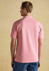 Gaiši rozā polo krekls ar mīkstu tekstūru, īsām piedurknēm un ribotu apkakli, kombinēts ar zilajiem šortiem, kuriem ir mazi izšūti raksti.