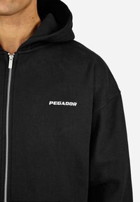 Casaco de capuz preto com fecho de correr, feito de tecido macio. Apresenta um capuz com cordão e o logótipo branco "PEGADOR" no peito esquerdo. Detalhe de fecho de correr prateado.