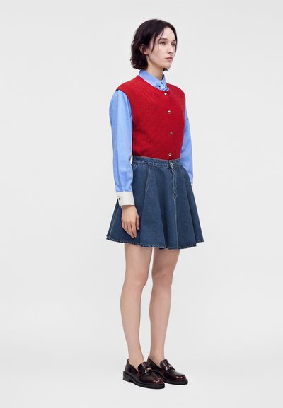 Femme portant un gilet rouge boutonné sur une chemise bleue, une jupe en jean et des mocassins foncés, debout devant un fond clair uni.