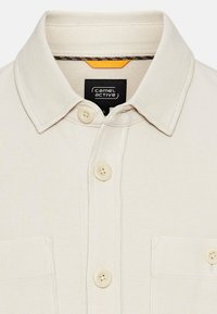 Beige skjorte med krave lavet af tekstureret stof, med to knapper, en brystlomme og et indre mærke med "camel active" branding.