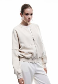 Lys beige zip-up sweatshirt med en afslappet pasform, ribbede manchetter og talje, parret med hvide bukser; glat, blød stoftekstur.