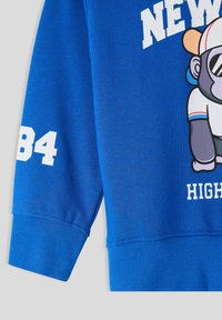 Felpa blu con polsini e orlo a coste, caratterizzata da una grafica di un monaco cartone e dal testo bianco "NEW YORK" e dal numero "84".