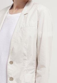 Blazer beige chiaro realizzato in tessuto morbido, con chiusura a un bottone, tasca sul petto e due tasche frontali. Sotto, un top bianco testurizzato.