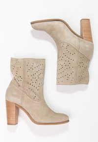 Botas de tobillo de ante beige con punta redondeada, tacón de madera apilada y detalles perforados en los lados. Textura suave, diseño casual.