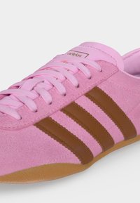 Ροζ αθλητικό παπούτσι Adidas με καφέ ρίγες, ροζ κορδόνια και λαστιχένια σόλα, που δείχνει το πάνω μπροστινό μέρος και τις πλευρικές λεπτομέρειες.
