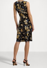 Mujer vistiendo un vestido negro sin mangas, a la altura de la rodilla, con estampado floral en amarillo, blanco y azul, combinado con sandalias de tacón alto negras.