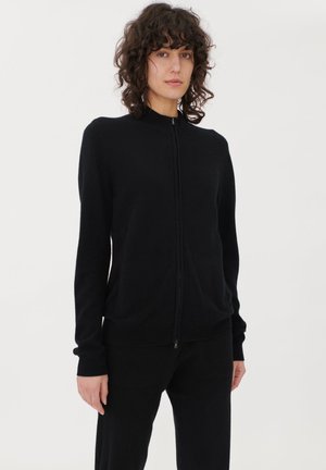 Frau mit lockigem Haar, die einen schwarzen Reißverschluss-Pullover und schwarze Hose trägt, steht vor einem schlichten hellen Hintergrund.