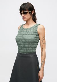Débardeur matelassé vert avec un motif texturé en forme de losanges, associé à une jupe sombre. Le mannequin porte des lunettes de soleil courbées de couleur claire.