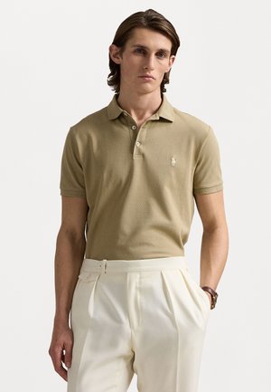 Beige poloshirt met een zachte textuur en een drukknoopsluiting met drie knopen, met een klein logo op de linkerkant van de borst. Gepaard met crèmekleurige broek.