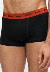 Gros plan d’un torse masculin portant un boxer noir avec une ceinture rouge Diesel, montrant la peau naturelle et les poils sur les jambes et le bas de l’abdomen.
