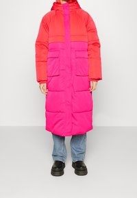 Manteau long rembourré avec capuche, présentant un design en bloc de couleurs rouge et rose. Comprend deux poches avant et des poignets côtelés.