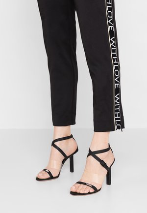 Černé sandály na podpatku s průhlednými popruhy a detailem s přezkou, na modelce v černých kalhotách s bílým logem.