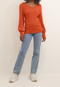 Maglione arancione a coste con scollatura ampia, maniche lunghe e polsini aderenti, abbinato a jeans dritti blu chiaro e sneakers bianche.