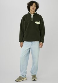 Jeune homme aux cheveux bouclés portant une polaire à fermeture éclair partielle vert foncé, un jean bleu clair et des baskets vert olive, debout devant un fond uni.