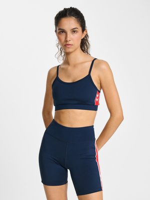 Vrouw met krullend haar in een navyblauwe sport-bh en bijpassende high-waisted wielrennersshorts met rode en witte strepen aan de zijkant, staand met handen achter de rug.