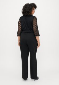 Svart jumpsuit med spetsöverdel, trekvartsärmar, avslappnad passform och vida byxor, kombinerad med svarta klackar.