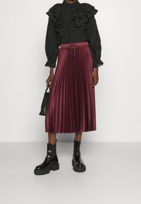 Blusa nera con volant a maniche lunghe, gonna midi plissettata bordeaux con vita regolabile, abbinata a stivali alla caviglia neri chunky e una borsa nera.