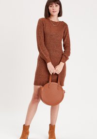 Vestido de suéter marrón de punto con mangas largas, escote en V y patrón texturizado. Combinado con un bolso circular marrón y botas de tobillo.