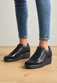 Baskets compensées noires en cuir et daim, avec lacets, fermeture éclair latérale et texture lisse et brillante.