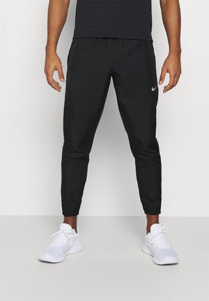 Mężczyzna ubrany w czarne spodnie dresowe Nike i białe buty do biegania Nike stoi na tle jednolitej białej ściany.