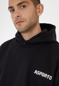Sudadera de algodón negra con cuello alto y cierre de botones, decorada con un logo impreso en blanco "ASPORTO" en la parte delantera. Textura suave.