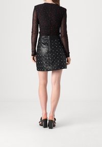 Femme portant un haut noir transparent à manches longues avec des pois rouges, une mini-jupe noire avec des clous argentés et des sandales noires à talons, vue de dos.