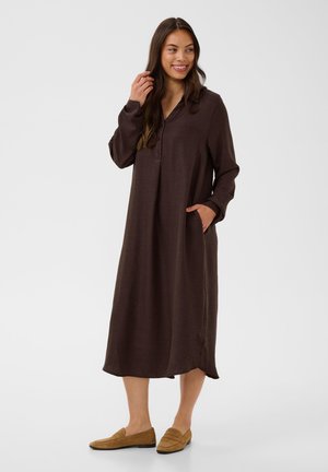 Vrouw draagt een lange donkerbruine jurk met knopen en lange mouwen en beige loafers, glimlacht en poseert met één hand dicht bij haar haar.
