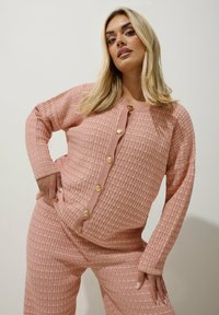 Rosa stickad cardigan med ett texturerat mönster, prydd med guldfärgad knappar och ribbade ärmslut. Matchas med tillhörande byxor.